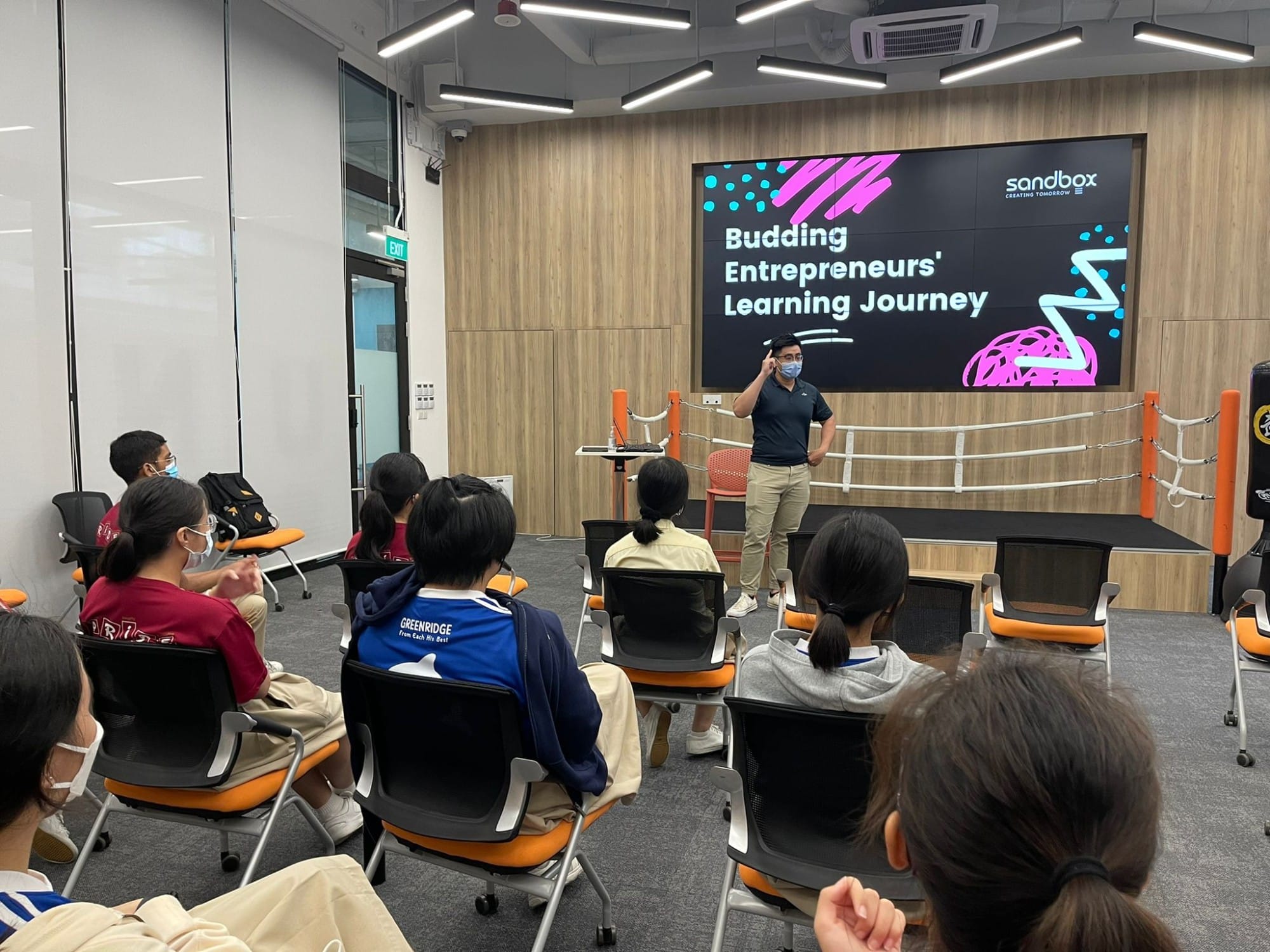A masked presenter speaks to seated students in front of a screen reading Budding Entrepreneurs Learning Journey at Sandbox.